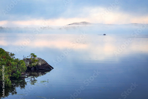 small fishing boat piercing through the fog on a calm lake with the hills in the background during sunrise in the country.