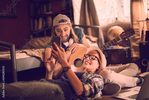 Young couple using a smartphone while playing the guitar in the bedroom at home