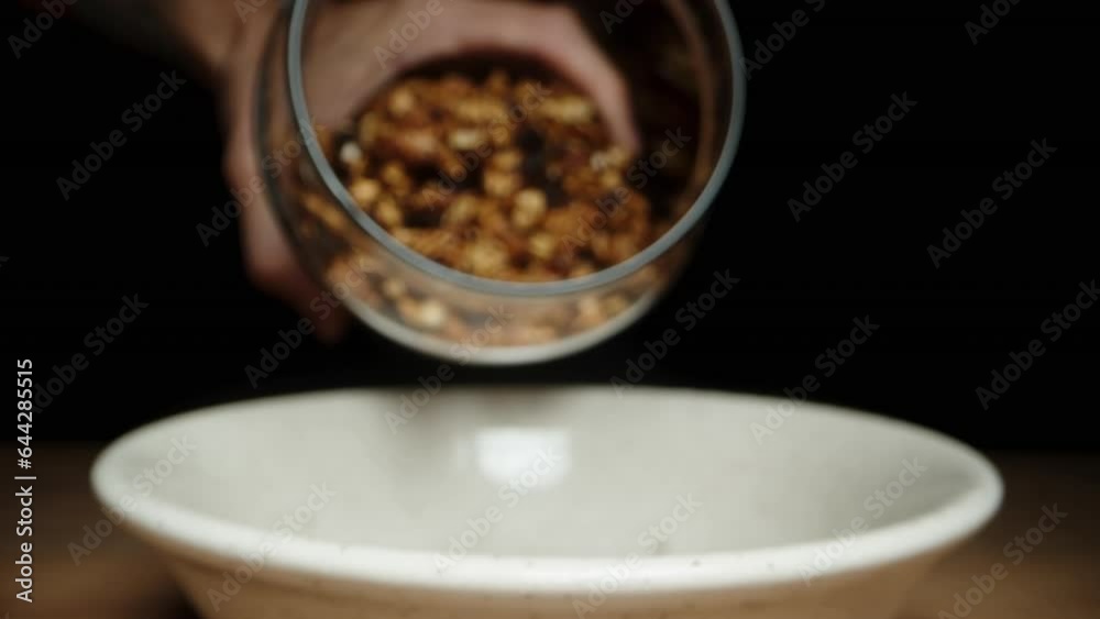 Against a black background, I pour granola from the jar onto the plate while the camera moves forward, in slow motion. close up Against a black background, I pour granola from the jar onto the plate while the camera moves forward, in slow motion. close up
