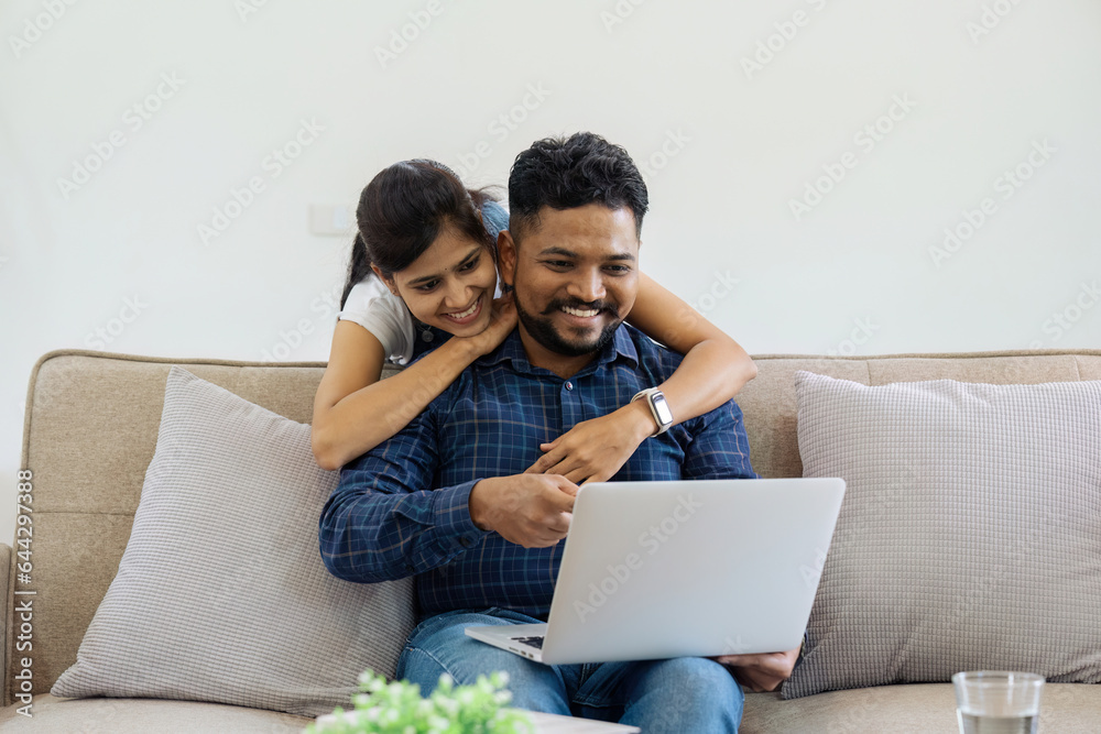 Happy loving family Indian couple hugging on couch at home and focused on laptop screen watching romantic movie