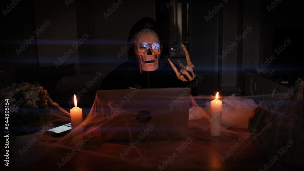 Mysterious man in scary skeleton costume show rocker symbol hand sitting table of laptop with cobwebs and candles in dark room, vignette. Advertising scary game.