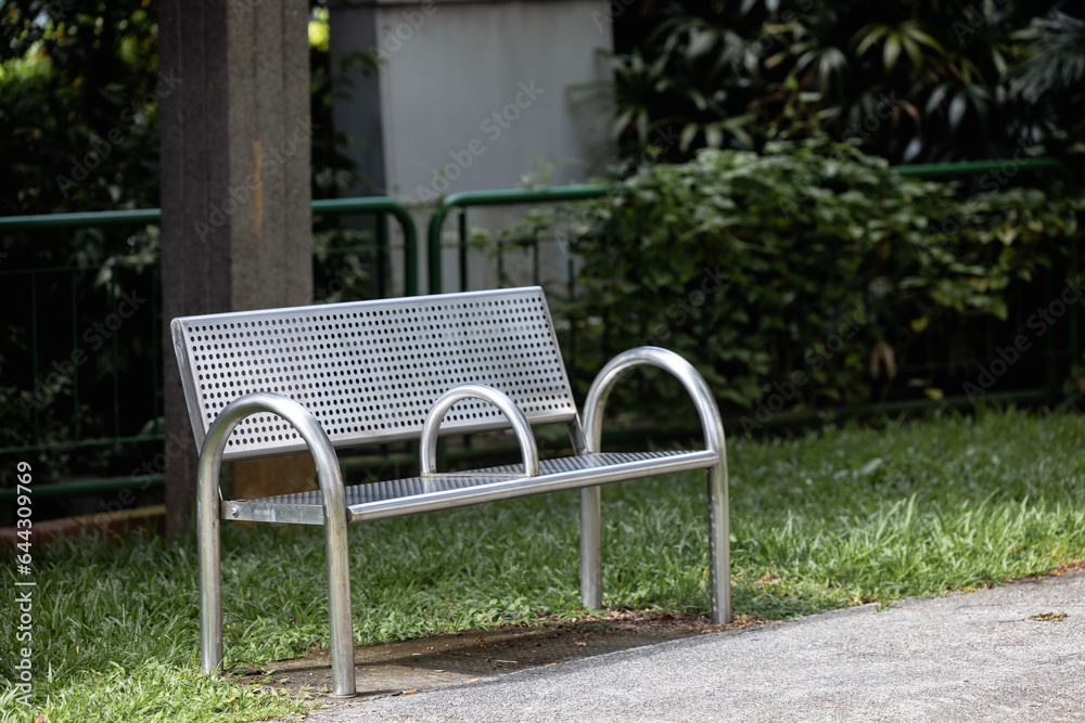 A metal bench stands in the park on the street, there are no people around, behind the bench you can see a green fence and a tall pole