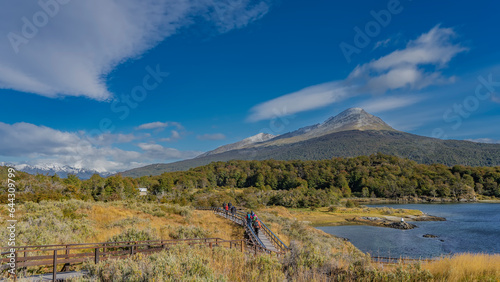 The wooden footpath is laid in the Tierra del Fuego National Park. Tourists walk along the path near the shore of the blue lake. Green forest in the distance. A mountain against a blue sky, clouds. 