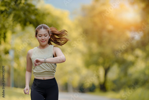 Fototapeta Naklejka Na Ścianę i Meble -  Beautiful Asian female jogger running in the park and looking at her watch to see her running timing in the park background.