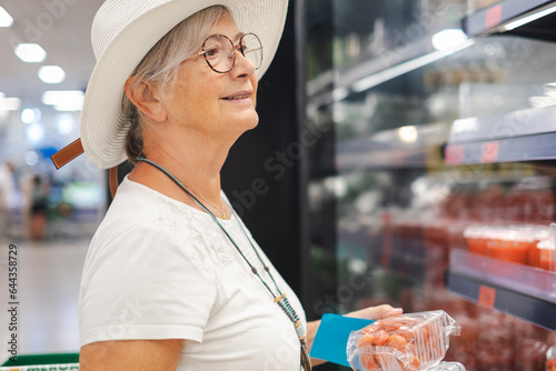 Smiling attractive senior customer woman with hat selecting tomatoes at supermarket. The elderly pensioner pays attention to the increase in prices