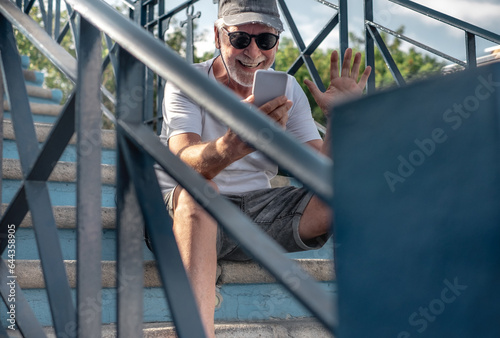 Video call concept. Smiling handsome bearded man in hat and sunglasses using online communication by phone sitting on outdoor staircase. Elderly male enjoying free time, tech and social