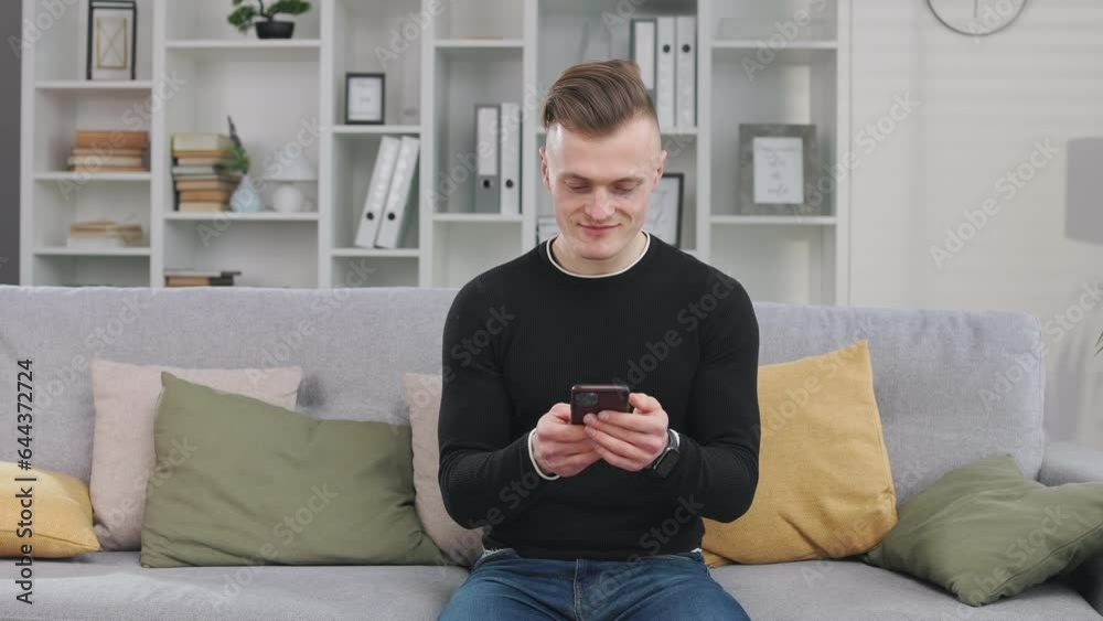 Front view of handsome blonde man with modern hairstyle sitting on couch and looking at smartphone. Young gentleman dressed in jeans and black sweatshirt chatting with girlfriend using mobile apps. Front view of handsome blonde man with modern hairstyle sitting on couch and looking at smartphone. Young gentleman dressed in jeans and black sweatshirt chatting with girlfriend using mobile apps.