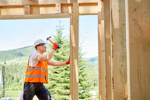 Side view of professional worker, builder standing, holding hammer and wooden gilder, beating. Man wearing vest and helmet, building wooden house in forest. Concept of building.