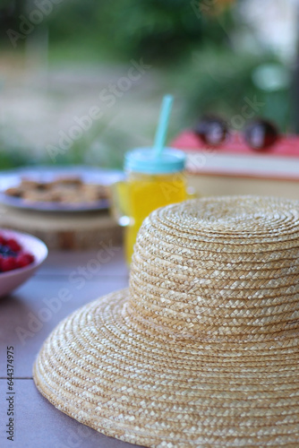 Straw hat, orange soda in the cactus shaped cup, bowl of fresh blueberreis and raspberries, plate of chocolate chip cookies, stack of books and sunglasses in the garden. Selective focus.