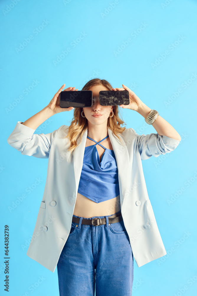 Social media, influencer. Young girl holding two mobile phones on eyes against blue studio background. Concept of human emotions, youth, fashion, lifestyle. Copy space for ad Social media, influencer. Young girl holding two mobile phones on eyes against blue studio background. Concept of human emotions, youth, fashion, lifestyle. Copy space for ad