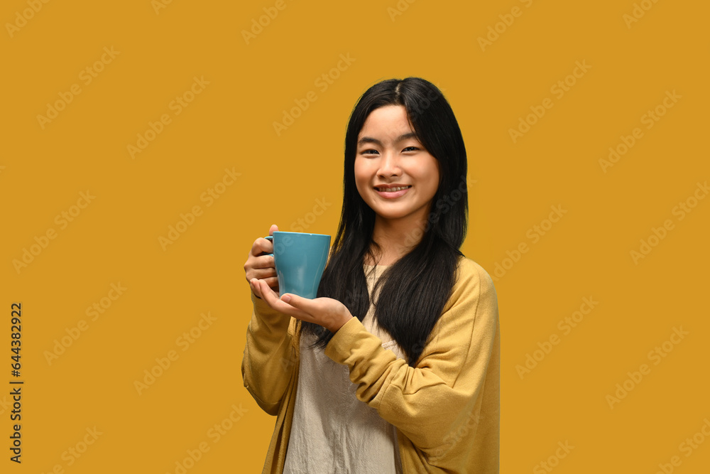 Happy teenager girl hold a ceramic mug of hot drink isolated over yellow background