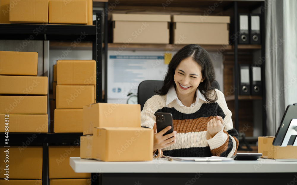 Happy young Asian woman entrepreneur, Smile for sales success after checking order from online shopping store in a smartphone at home, Concept of merchant business online and eCommerce. Happy young Asian woman entrepreneur, Smile for sales success after checking order from online shopping store in a smartphone at home, Concept of merchant business online and eCommerce.