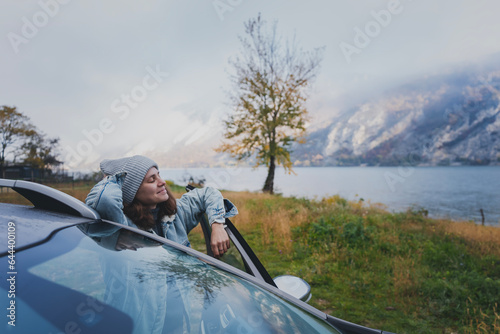 Young woman traveling by car standing against the backdrop of a beautiful mountain landscape with a lake