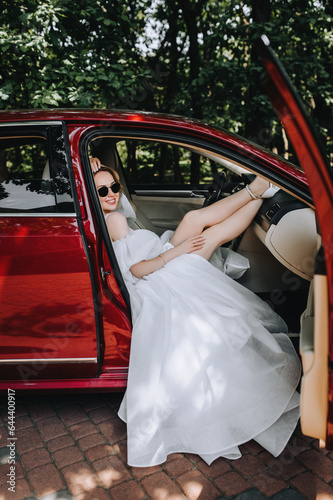 A beautiful blonde bride with a bouquet in sunglasses sits in a red car. Wedding photography, portrait, lifestyle. © shchus