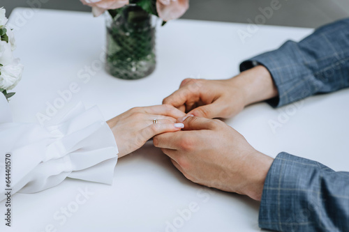 The bride and groom are sitting at the table of a cafe, a restaurant with a glass vase of flowers and holding hands with golden rings on their fingers. Wedding photography, portrait.