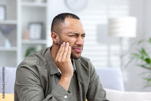 Close-up photo of a young African-American man suffering from a severe toothache at home on the couch. He holds his hand to his cheek, grimacing in pain