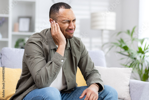 Severe pain and inflammation in the ear of a young African American man sitting on the couch at home and holding his head © Tetiana