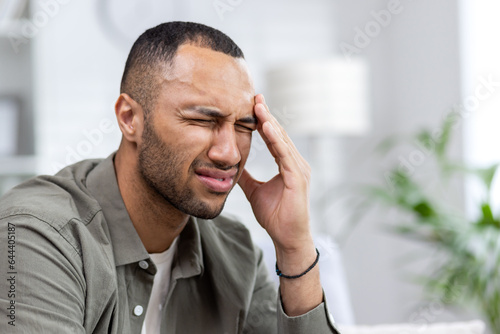 Close-up photo of a young African American man suffering from a severe headache. Sitting at home on the sofa, grimacing in pain, holding his head with his hand, doing a massage