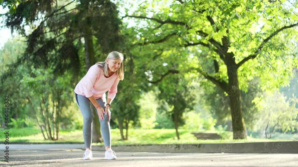 Senior gray haired woman suffers from painful severe knee joint ache during a run in city park. Mature old female massaging stretching trauma injury while jogging outdoors. Leg muscle cramp calf sport