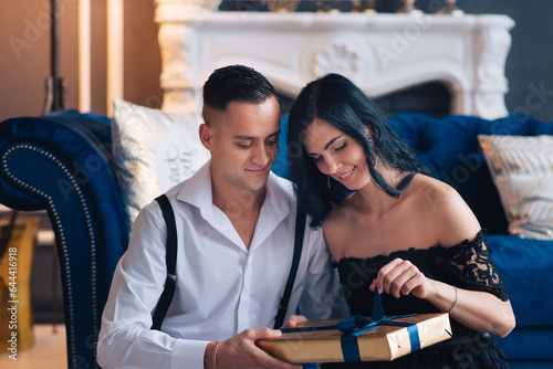 Portrait of a young couple in their living room at christmas eve. Lovely couple in love embracing in a room in a living room with a blue interior and a lamp on. Couple opens a gift.