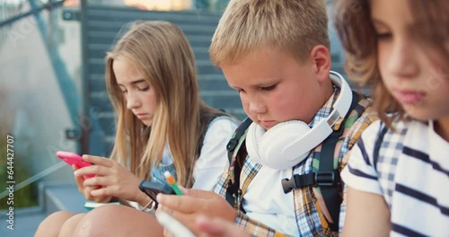 Close up of group of schoolchildren sitting on stairs outdoors using mobile phone communicating online. Diverse kids use internet on cellphone while relaxing on street at sunny day