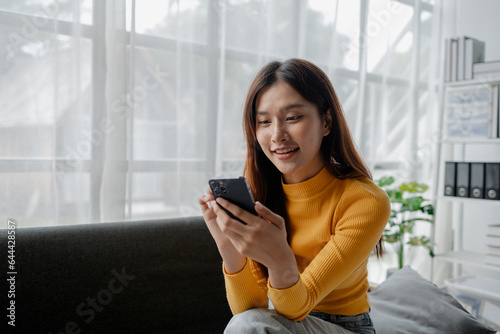 Asian woman is in the living room and uses social media on a smartphone. Female company employee on vacation. Using vacation time and free time to relax at home. Weekend vacation day doing activities. #644428587