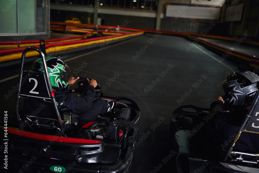 two multicultural men in helmets and sportswear driving go kart on indoor circuit, competitors