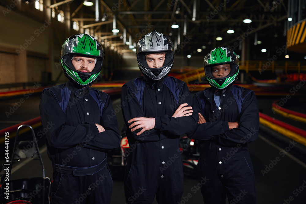 three confident go kart drivers in sportswear and helmets standing with folded arms on circuit three confident go kart drivers in sportswear and helmets standing with folded arms on circuit