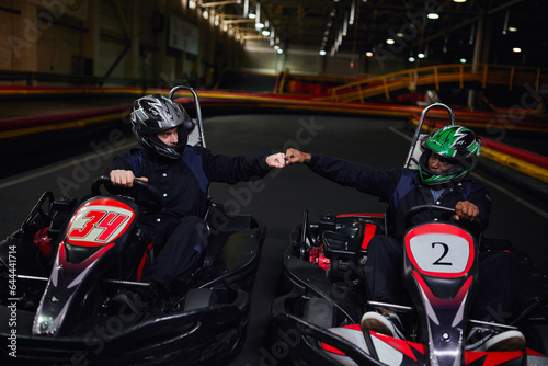 diverse go kart drivers in helmets fist bumping and sitting in sport cars for karting on circuit