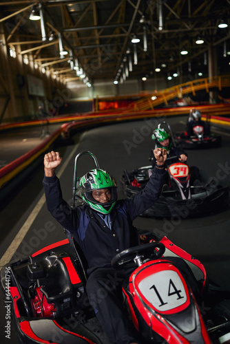 excited african american go cart driver in helmet raising hands and winning race on circuit, go-kart