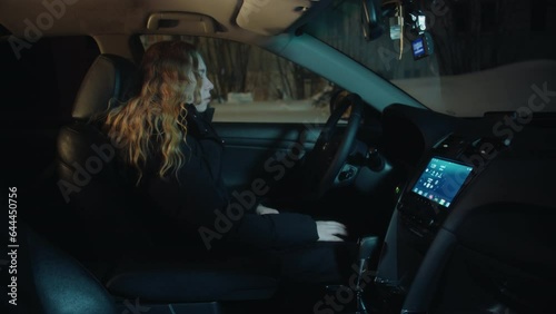 A tired asleep young woman with long curly hair and in a black jacket turns off the engine from the button, unfastens the belt and gets out of the car, closes the door and leaves at night in winter photo