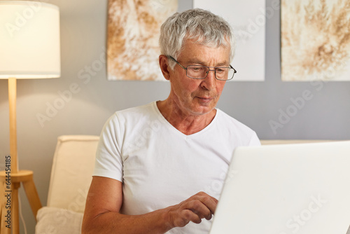Senior gray haired man wearing white T-shier and glasses sitting on couch in living room holding laptop typing on keyboard looking at display retired male working on online
