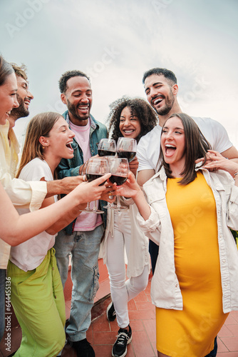 Vertical. Group of young cheerful friends smiling and toasting red wine glasses at rooftop birthday party. Happy adult people having fun clinking wineglasses at weekend celebration. Buddies with booze