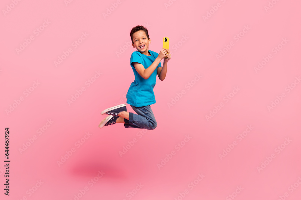 Full length photo of funky positive small boy wear blue t-shirt texting modern device jumping high isolated pink color background