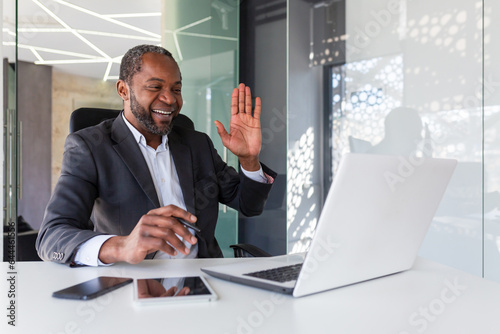 Joyful successful african american man inside office at workplace talking remotely on video call, man smiling and looking at laptop screen, waving hand in greeting gesture.