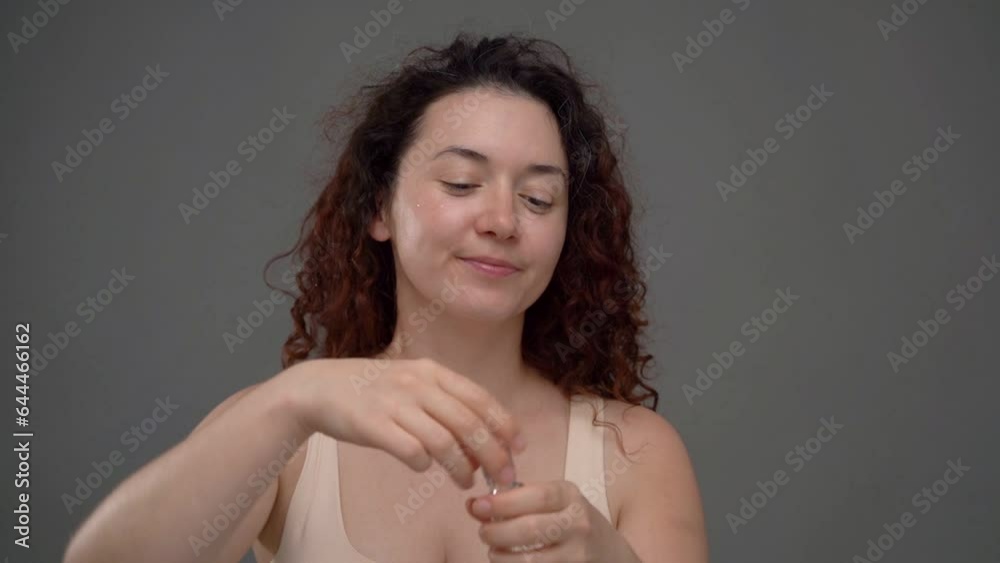 Skin care face massage routine with serum video footage. Beautiful 30 years old woman applying cosmetics. Curly haired Caucasian woman at gray background studio shot. Smiling positive emotions Skin care face massage routine with serum video footage. Beautiful 30 years old woman applying cosmetics. Curly haired Caucasian woman at gray background studio shot. Smiling positive emotions