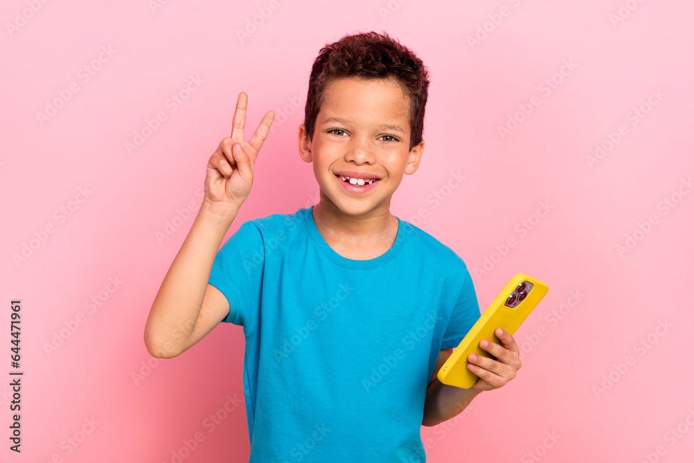 Photo portrait of charming small boy hold telephone showing v-sign dressed stylish blue garment isolated on pink color background Photo portrait of charming small boy hold telephone showing v-sign dressed stylish blue garment isolated on pink color background
