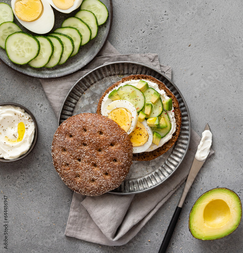Rye bread sandwiches with cream cheese spread, boiled eggs, avocado cubes and sliced fresh cucumber. Healthy eating concept. Delicious breakfast, snack or lunch. Top view, grey background. 