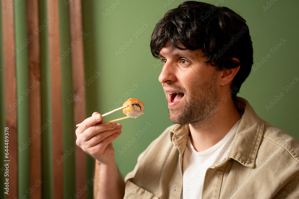 Fototapeta premium Young handsome man wearing beige shirt eating sushi roll with salmon using chopsticks. 