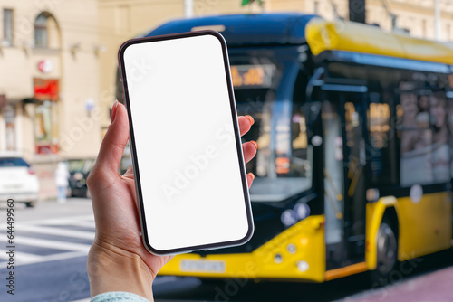 Girl holds a mock-up of a smartphone with a white screen in her hands at a bus stop against the backdrop of a bus.