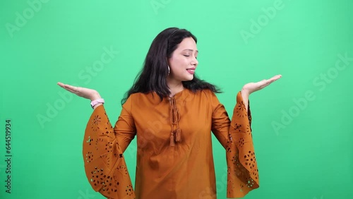 Beautiful Young indian woman casually dressed with empty place on her palm showing invisible product and choosing the right one by comparing it over removable green chroma background.