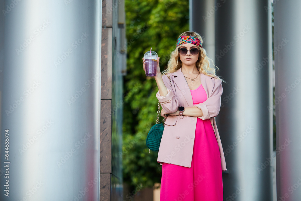 View from stylish female model with cup of drink on hand wears trendy pink jacket and trousers in urban place. Fashion woman portrait. View from stylish female model with cup of drink on hand wears trendy pink jacket and trousers in urban place. Fashion woman portrait.