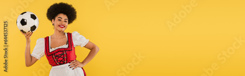 happy african american bavarian waitress posing with soccer ball and hand on hip on yellow, banner © LIGHTFIELD STUDIOS