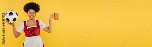happy african american bavarian waitress in dirndl with beer mug and soccer ball on yellow, banner © LIGHTFIELD STUDIOS