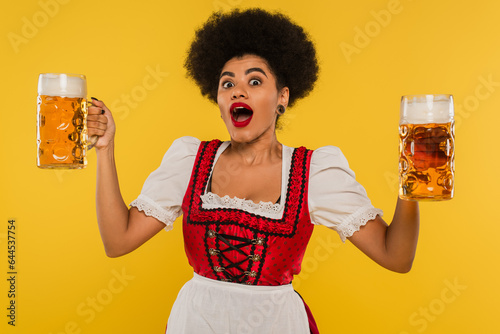 thrilled african american bavarian waitress with open mouth standing with full beer mags on yellow