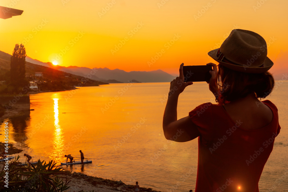 Fototapeta premium Silhouette of a woman taking a photo at sunset at Shkoder lake in Shiroka municipality, summer vacation. Albania