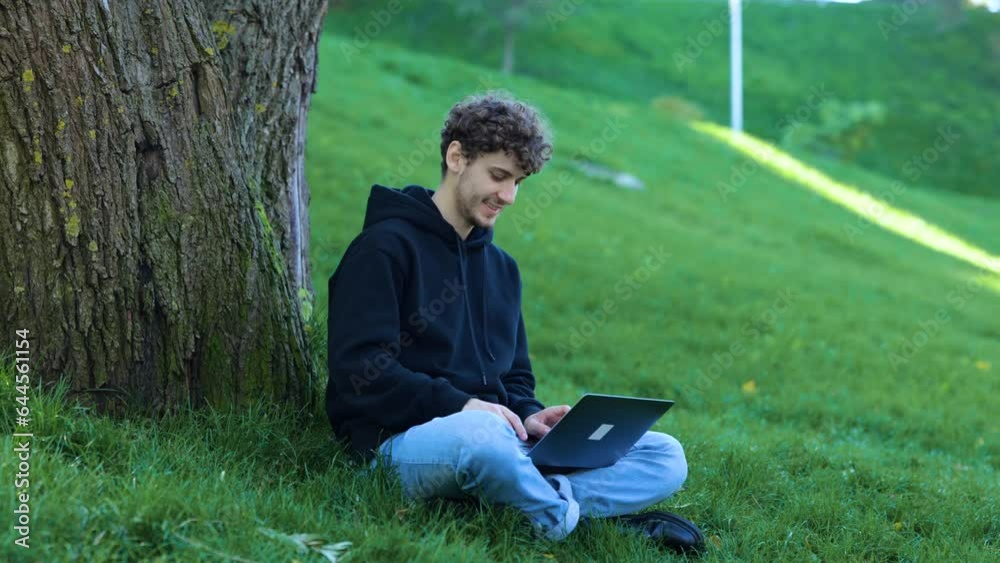Smiling guy using laptop while typing. Man sitting near the tree in the city park. Autumn season. Education, working concept. Slow motion Smiling guy using laptop while typing. Man sitting near the tree in the city park. Autumn season. Education, working concept. Slow motion