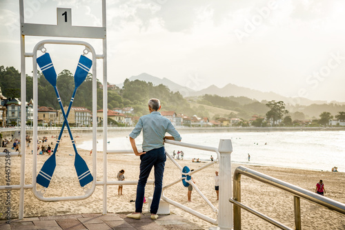 Elegant Elderly Man with His Back Looking at the Beautiful Sunset of Ribadesella Beach Leaning on the Railing of the Promenade with Oars at his side