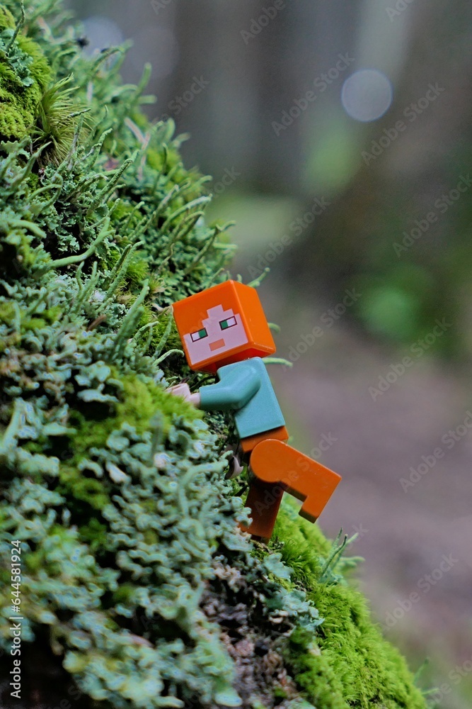 Obraz premium LEGO Minecraft figure of main character Alex climbing on forest rock densely covered with moss and lichen.