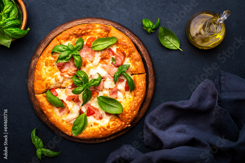 Homemade hot pizza with ham, mozzarella cheese, tomato sauce and green basil, black table background, top view